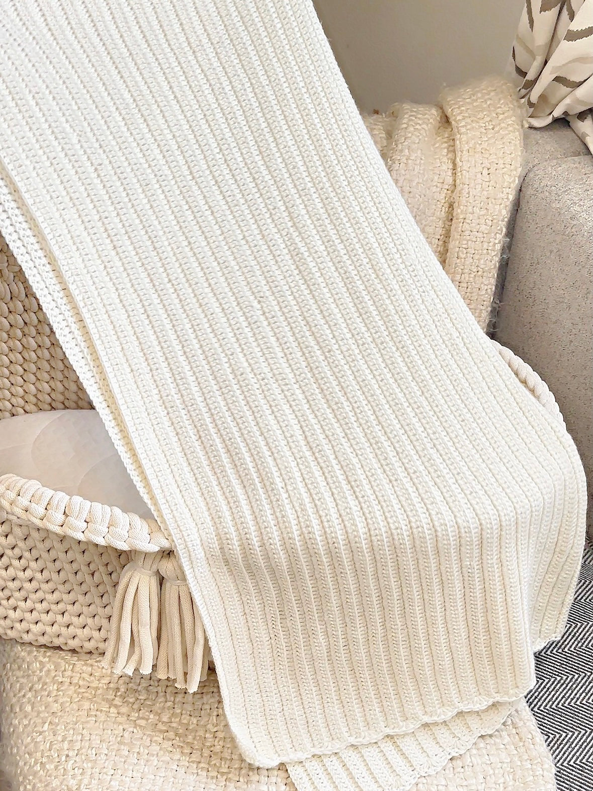 A close-up of a cream ribbed knit throw blanket draped over woven furniture, showing its soft texture and tasseled edge.