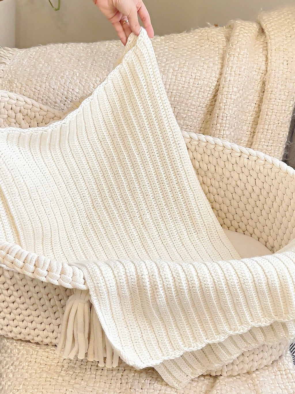 A cream-colored ribbed knit throw blanket with fringe tassels, draped over a woven basket with a hand holding up one corner.