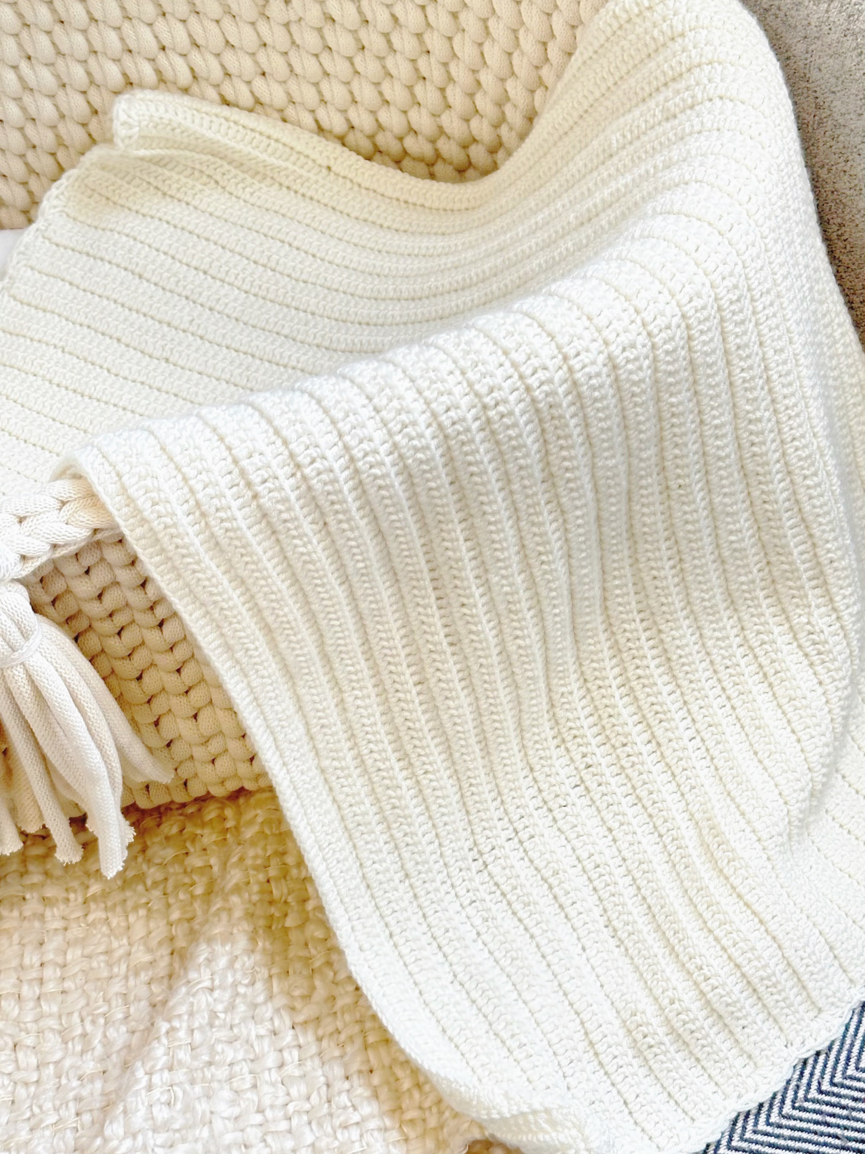 A close-up of a cream-colored ribbed knit blanket draped over a woven beige chair, showing its soft texture and tassels.