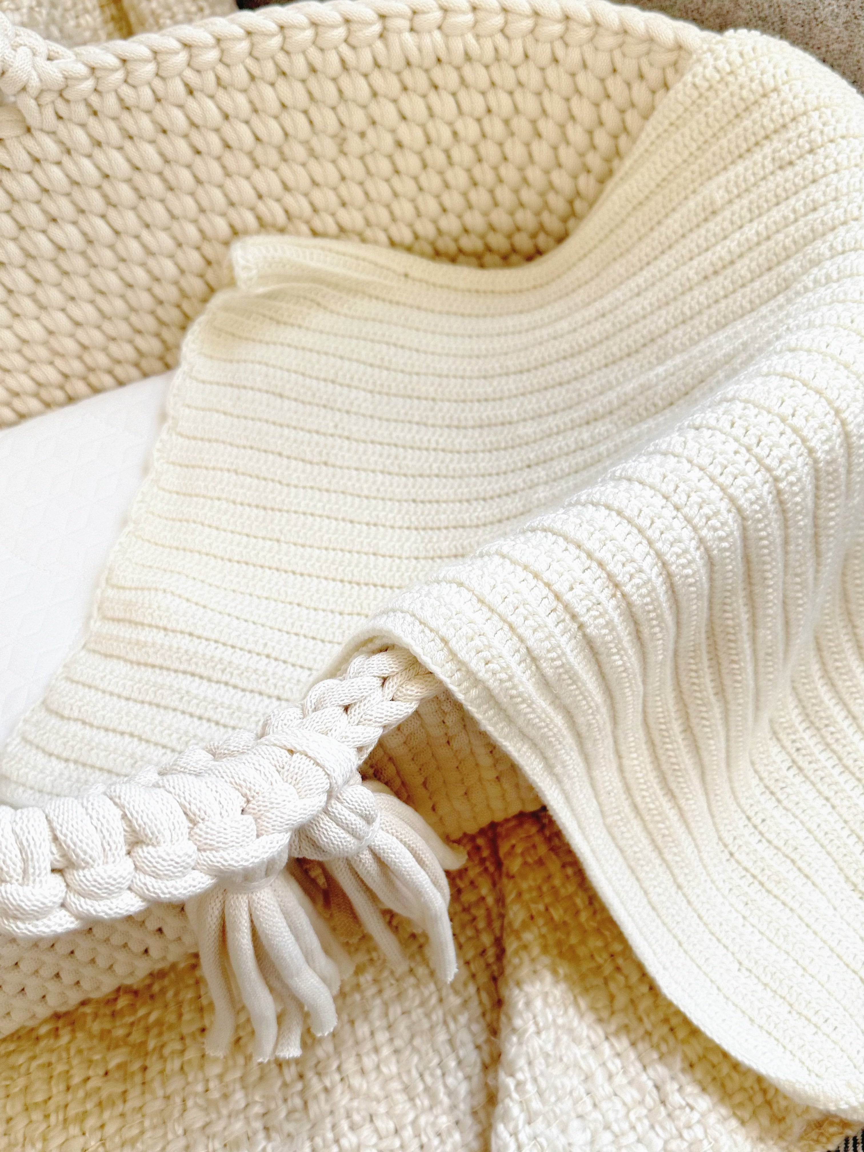 Close-up of a cream-colored knit blanket with a ribbed texture, braided edge, and tassels, draped over a woven basket.